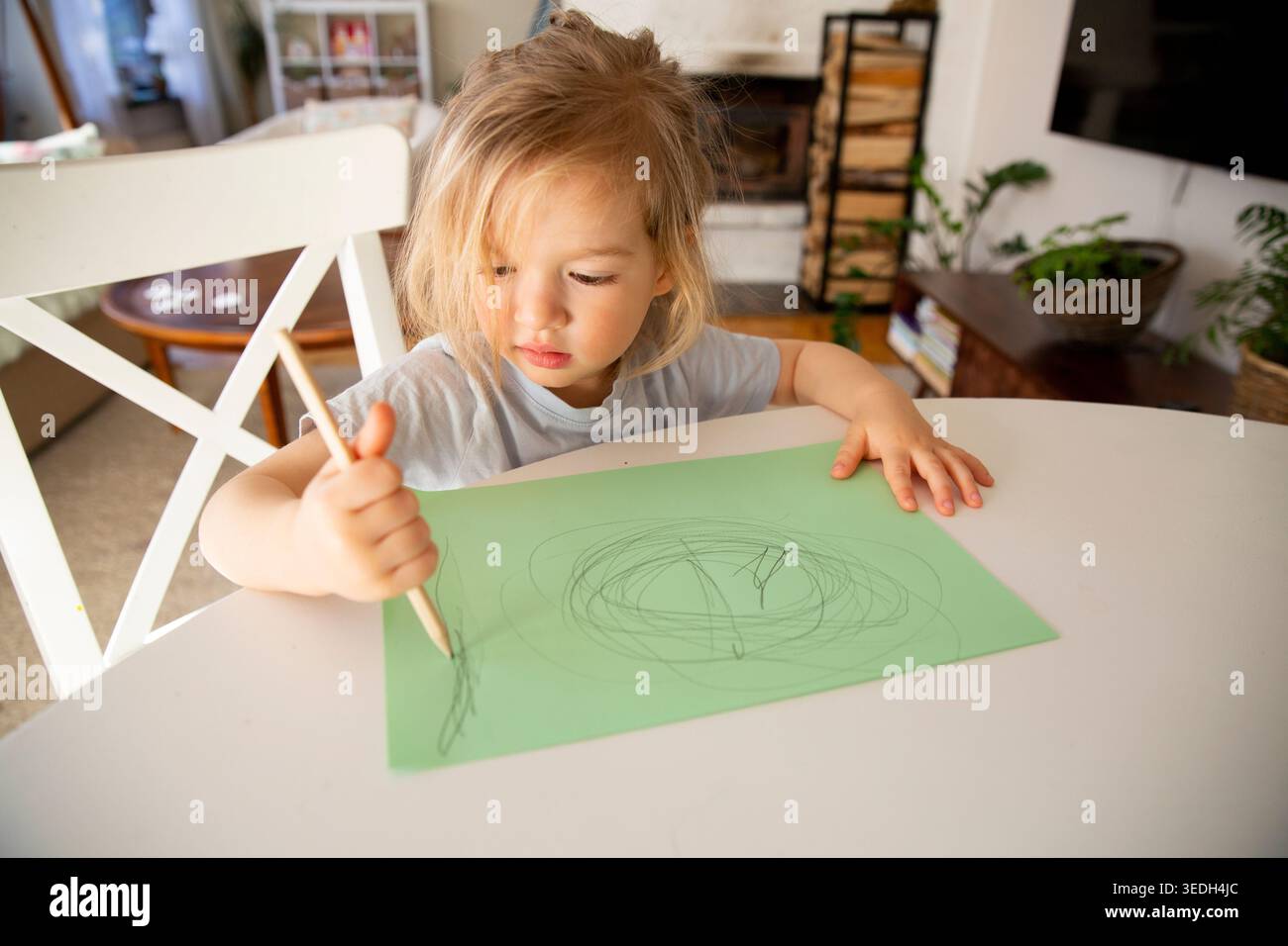 Enfant avec de longs cheveux dessinant des gribouilles sur papier vert avec un crayon en bois, assis à table dans une maison confortable, développement précoce créatif et jeu analogique Banque D'Images