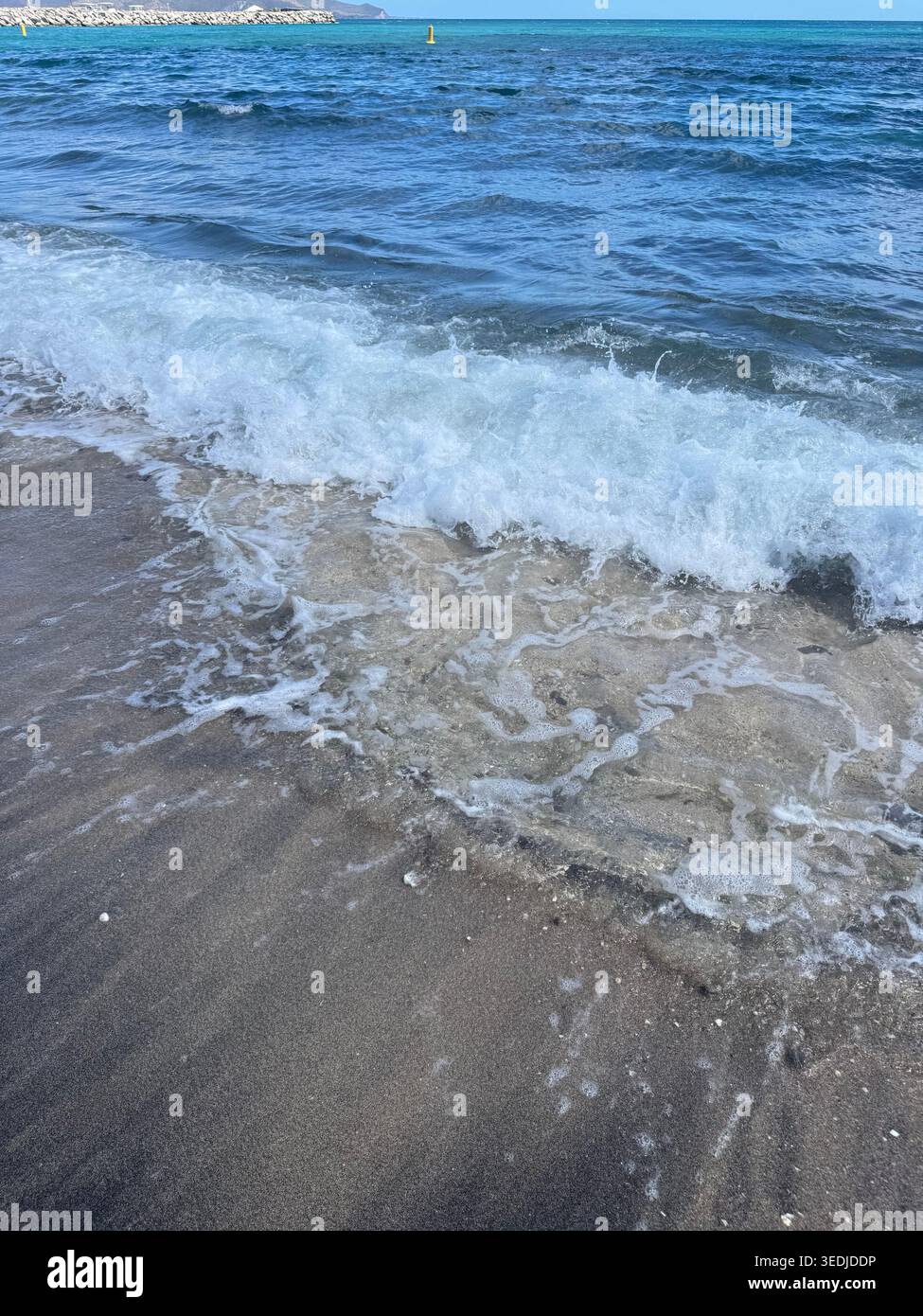 Gros plan de l'écume marine blanche et de l'eau qui reculent sur une plage de sable volcanique sombre et humide. - Image de stock capturée avec un smartphone