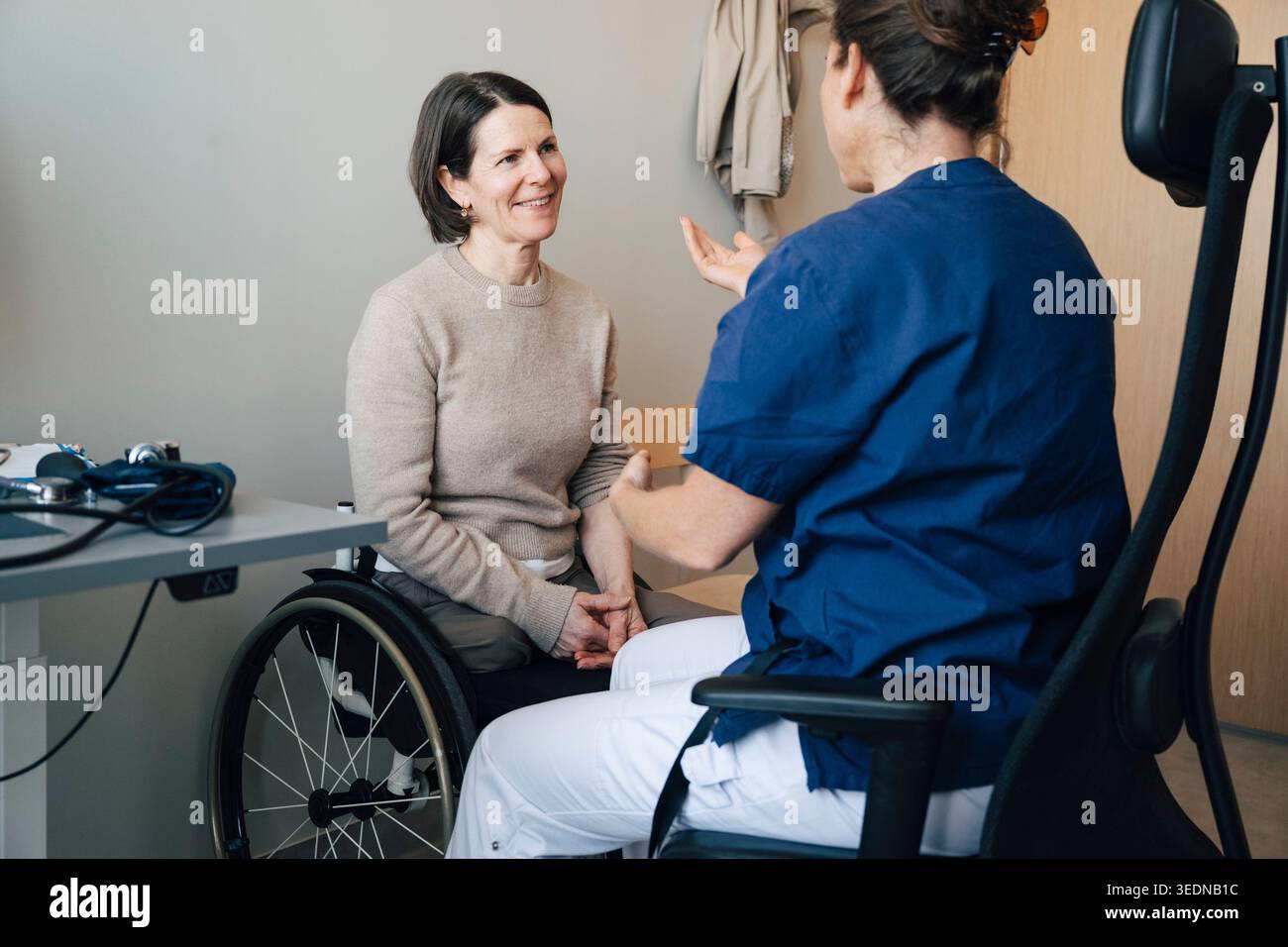 Femme mature souriante assise sur un fauteuil roulant écoutant le médecin féminin pendant l'examen médical dans la chambre d'hôpital Banque D'Images