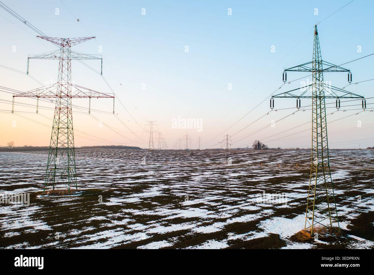 Des tours de transmission de puissance se dressent au-dessus d'un champ enneigé au crépuscule. L'horizon présente un doux dégradé de couleurs lorsque le soleil se couche, projetant une lumière douce sur le paysage hivernal. Banque D'Images
