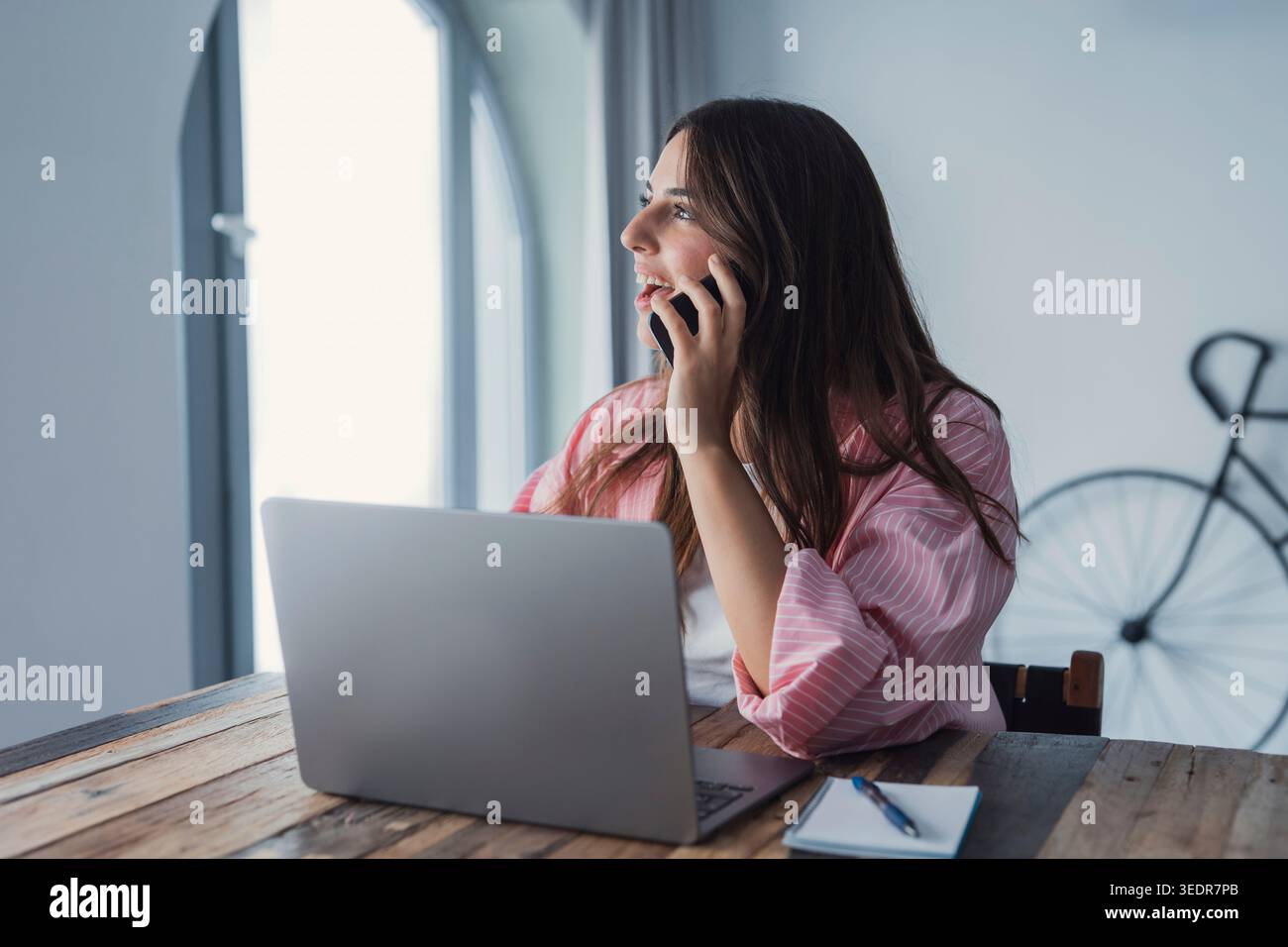 Une jeune femme professionnelle sourit en consultant un client au téléphone à son bureau, distrait de son ordinateur mais gardant un comportement amical et professionnel Banque D'Images