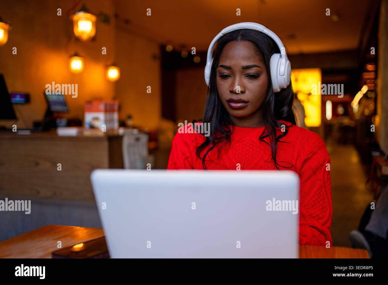 Jeune femme noire se concentrant sur un ordinateur portable, portant des écouteurs dans un café éclairé chaud Banque D'Images