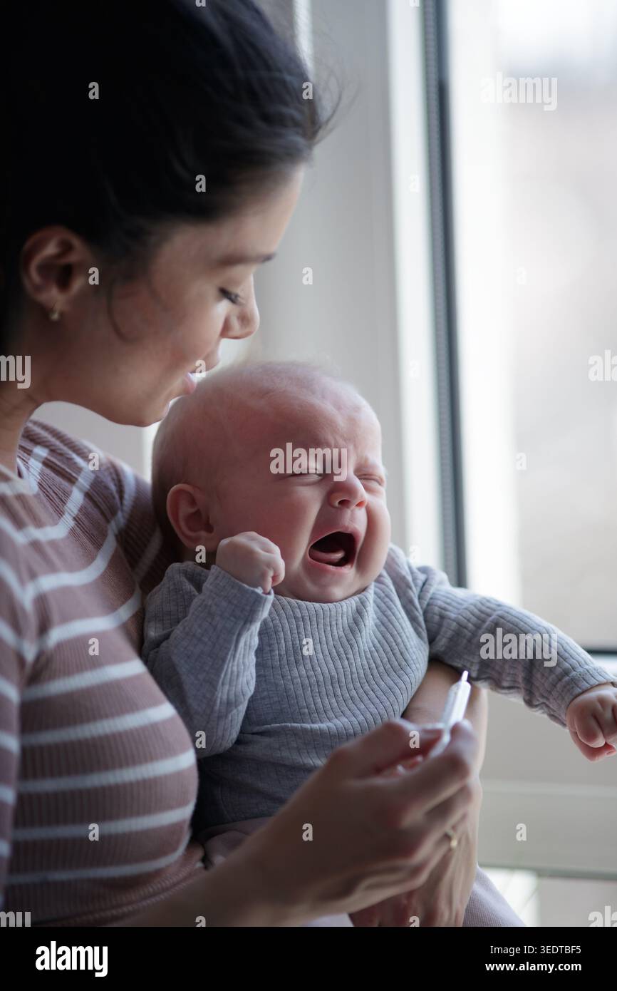 Une mère inquiète tenant le bébé pleurant dans une main à la maison et regardant le thermomètre pour la fièvre Banque D'Images