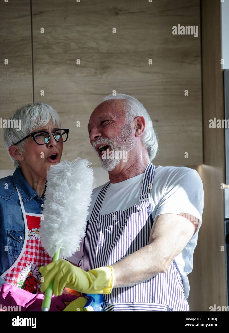 Couple mûr s'amusant tout en faisant des tâches ménagères dans la cuisine ensemble et en chantant avec Duster comme microphone Banque D'Images