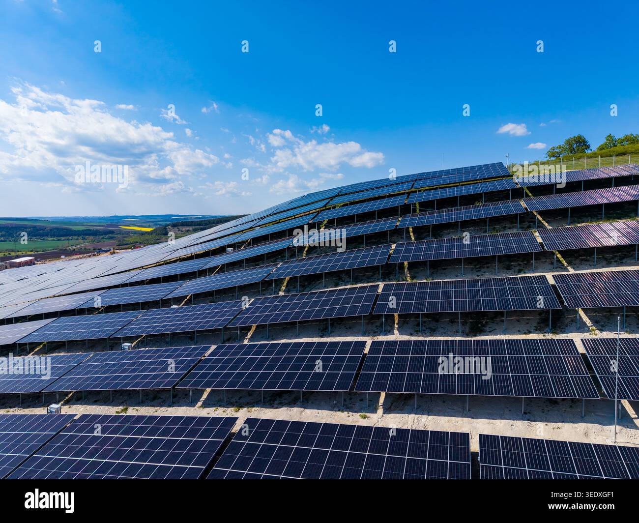 Des rangées de panneaux solaires bordent une zone inclinée, capturant la lumière du soleil pour l'énergie. Le paysage montre des champs et des arbres sous un ciel bleu avec peu de nuages. Banque D'Images