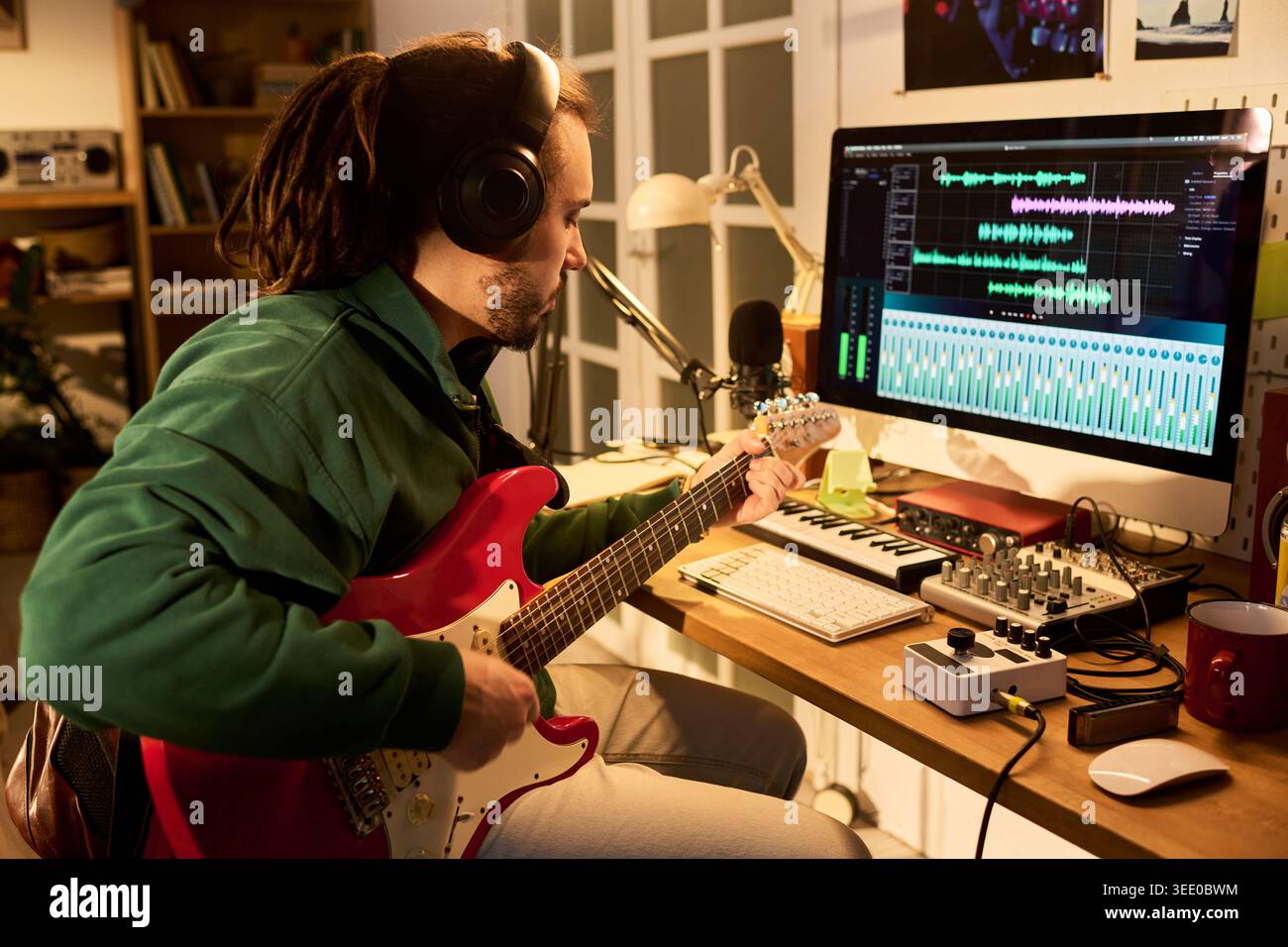 Jeune homme adulte enregistrant la piste de guitare électrique dans le studio de musique à domicile, surveillant le logiciel d'édition audio sur le bureau. Utile pour la création de contenu, podcast, marketing de production musicale Banque D'Images