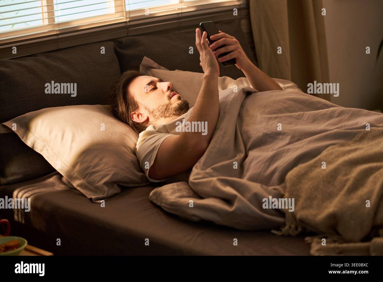 Jeune homme caucasien adulte allongé dans le lit et utilisant un smartphone la nuit, naviguant ou envoyant des messages avant de dormir. Concept pour la dépendance à l'écran, l'insomnie, le marketing digital de style de vie Banque D'Images