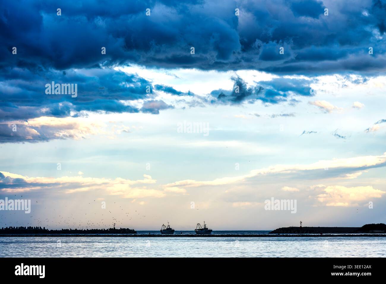Deux chalutiers de pêche entrant dans le canal du port à l'embouchure de la rivière Pescara, dans les Abruzzes, Italie, sous un ciel orageux avec des oiseaux de mer en vol. Banque D'Images