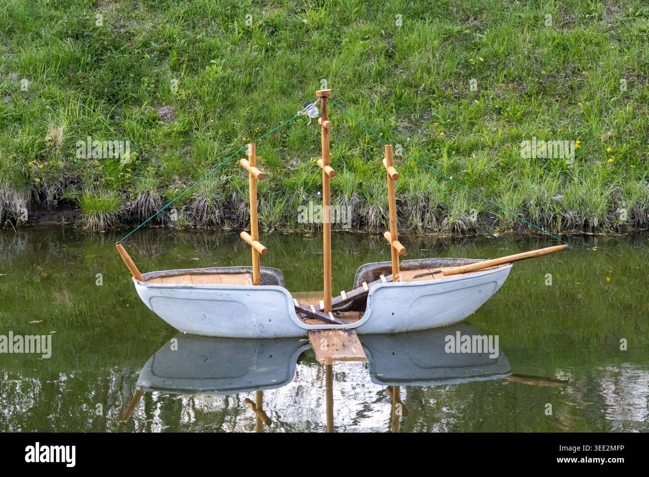 Un bateau en béton repose sur une eau calme, tenue par des poteaux en bois. De l'herbe et des plantes entourent la zone. La scène est lumineuse avec une lumière douce, montrant le beau rural Banque D'Images