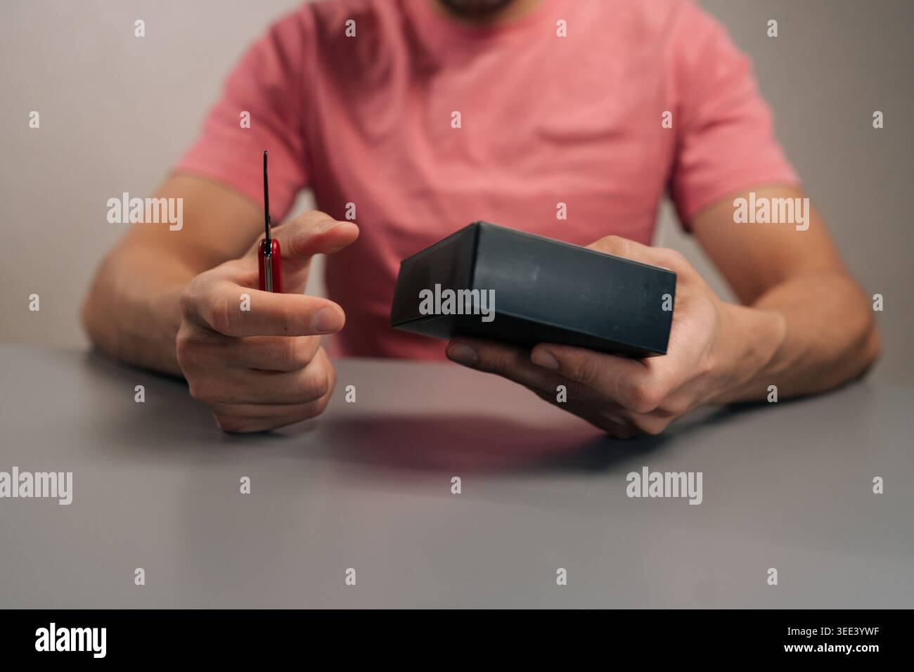 Mains de la personne utilisant le petit multi-outil rouge pour ouvrir, déballer et déballer le nouveau gadget de produit de la boîte d'emballage en carton noir assis à la table, révélant R Banque D'Images