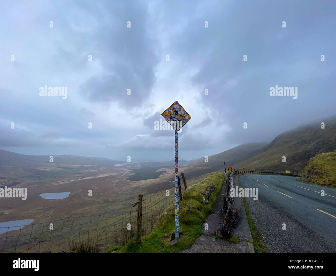 Panneau routier en diamant recouvert d'autocollants au sommet de Conor Pass avec vue aérienne panoramique sur la vallée de la péninsule de Dingle et les lacs, Kerry, Irlande Banque D'Images