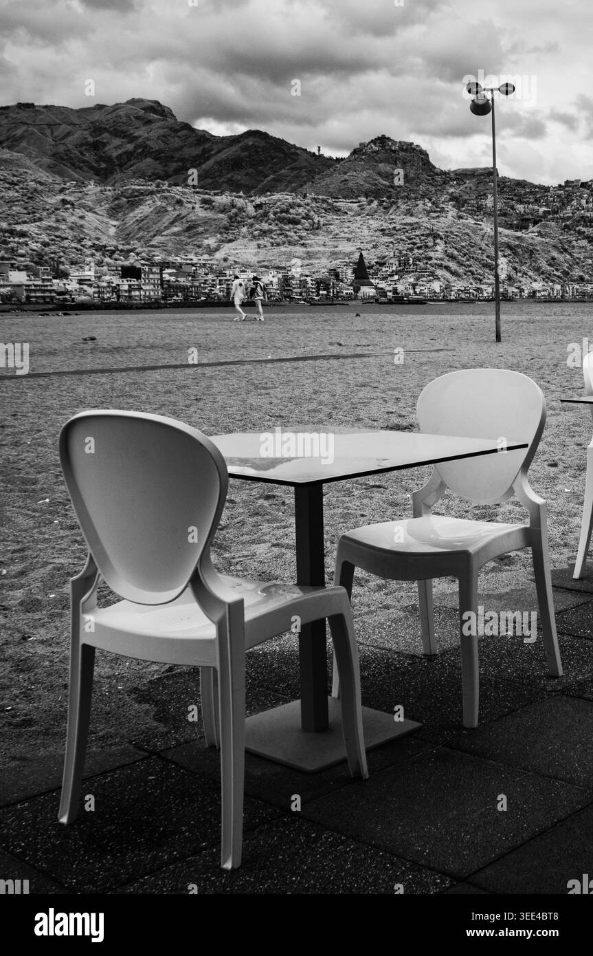 Photographie en noir et blanc capture une terrasse extérieure vide avec deux chaises et une table au premier plan. En arrière-plan, le littoral de Giardi Banque D'Images