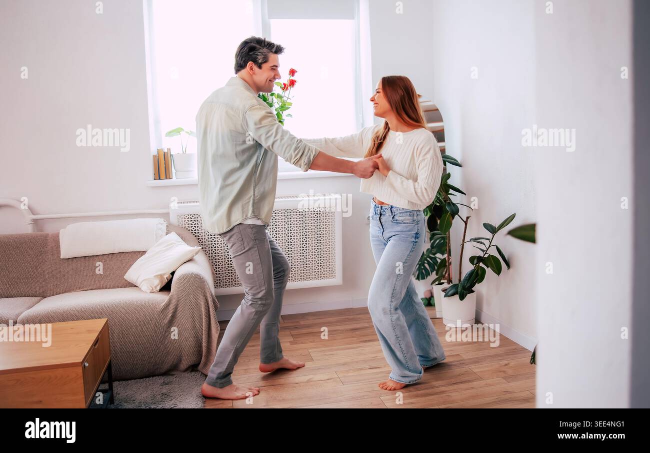 Heureux jeune couple dansant ensemble à la maison, homme romantique tenant des roses rouges et femme surprenante dans un salon moderne lumineux Banque D'Images