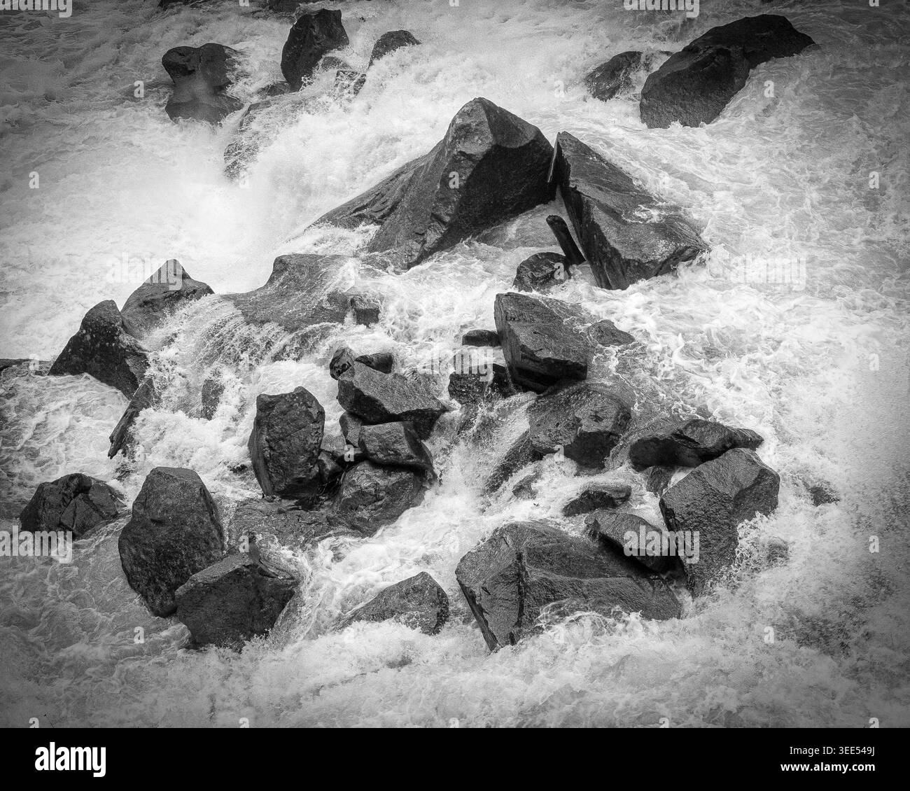 L'eau s'écrase contre de gros rochers le long du rivage par une journée nuageuse dans une rivière. Photographie en noir et blanc. Banque D'Images