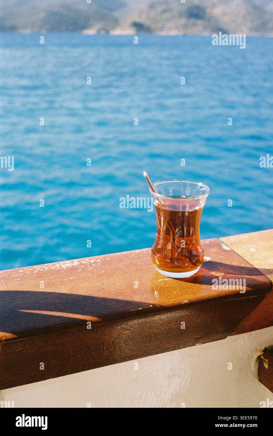 Verre de thé turc traditionnel avec thé noir sur une corniche en bois, surplombant une mer bleue vibrante et la côte montagneuse Banque D'Images