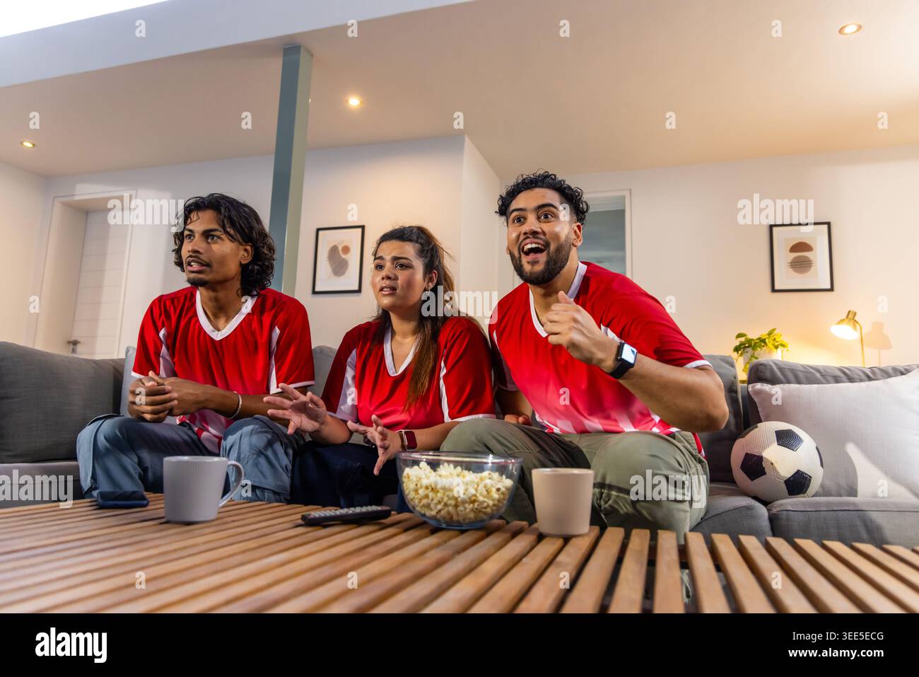 Divers amis regardant le football sur le canapé gris portant des maillots rouges à la maison avec pop-corn et ballon de football Banque D'Images