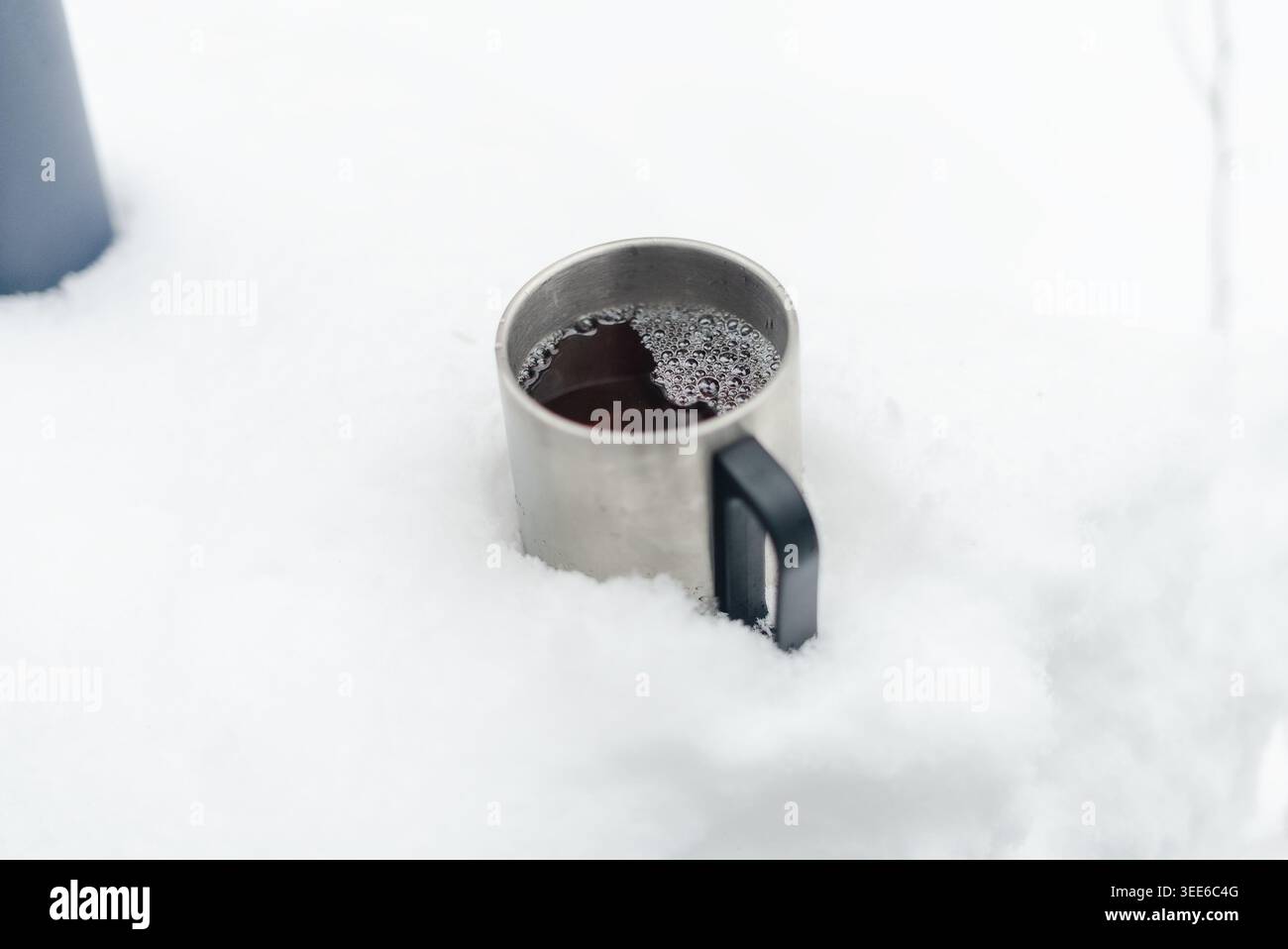 Tasse en métal rempli d'une boisson chaude sombre est en partie enfouie dans la neige fraîche à l'extérieur. La tasse en émail cuit doucement dans l'air froid de l'hiver. Un moment de boisson réchauffant lors d'une aventure par temps froid Banque D'Images
