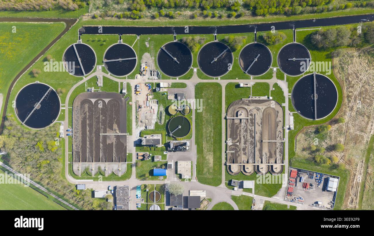 Vue aérienne de l'usine de traitement des eaux de RWZI - Geestmerambacht avec des réservoirs circulaires et des bassins rectangulaires entourés de champs verts et d'arbres dans le filet Banque D'Images