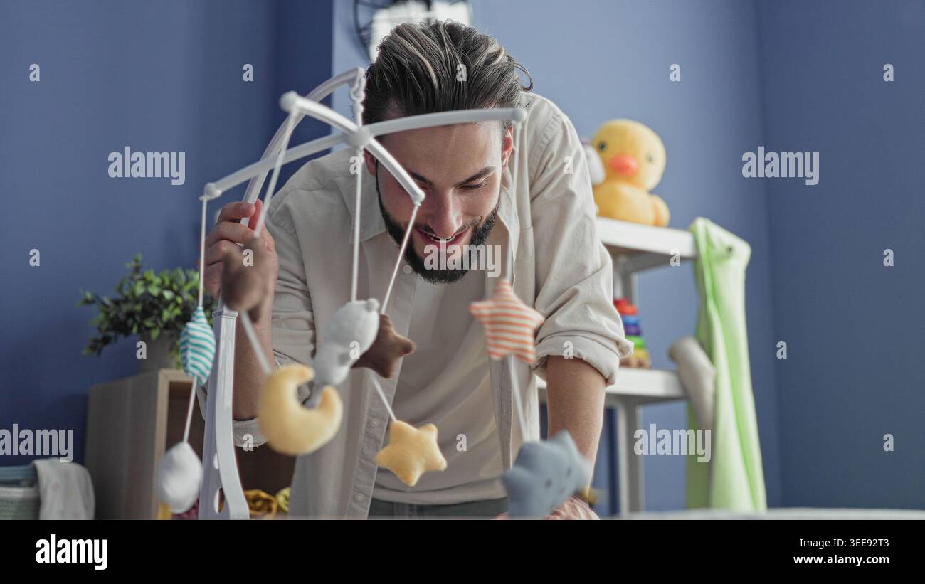 L'homme se penche sur le berceau ajustant un mobile suspendu tout en souriant dans une pépinière avec des jouets en peluche, un canard en caoutchouc et des anneaux empilables ; tendresse de liaison. Banque D'Images
