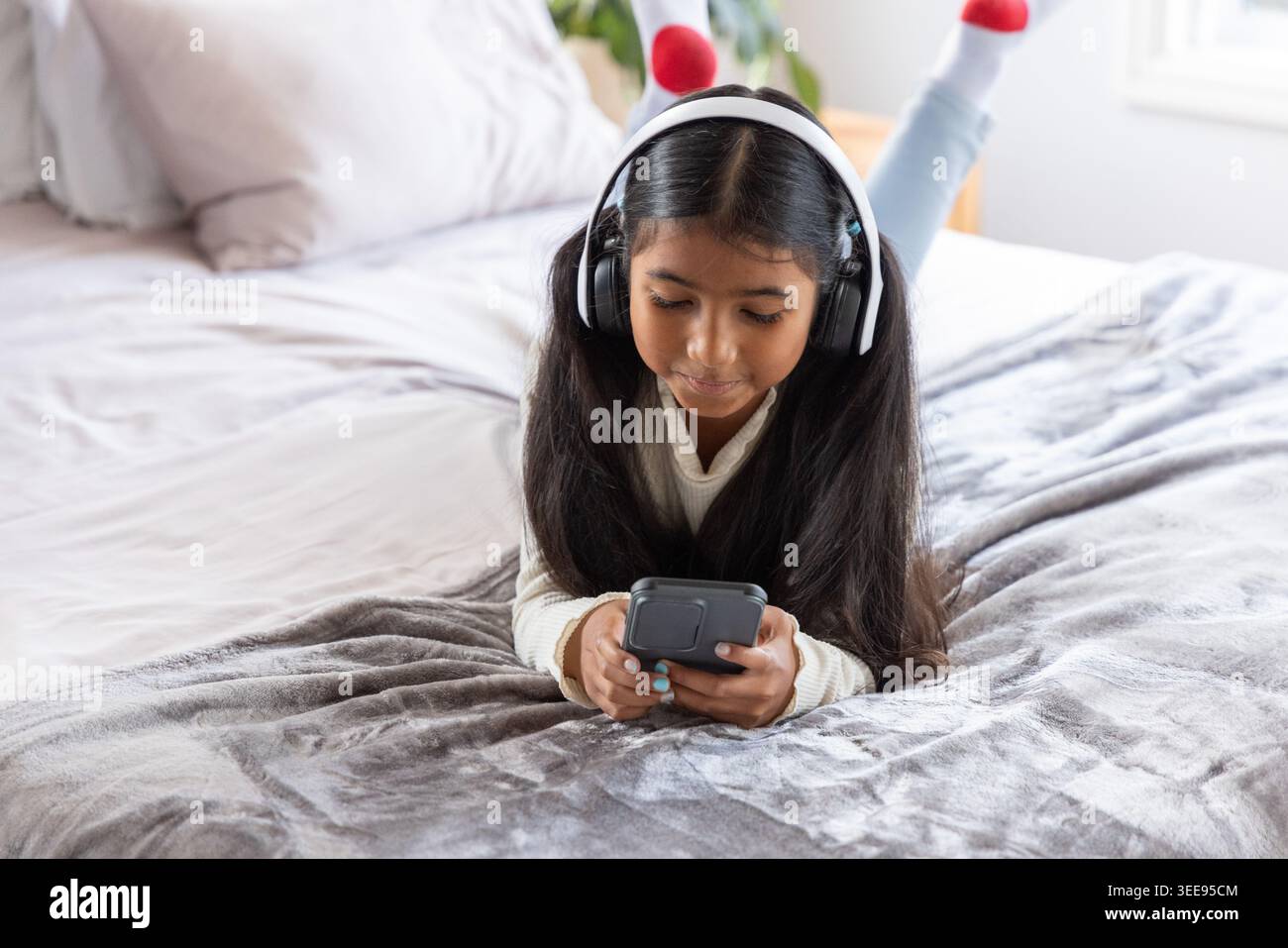 Fille indienne d'âge scolaire couchée sur le lit portant un pull tenant le téléphone et écoutant sur des écouteurs Banque D'Images