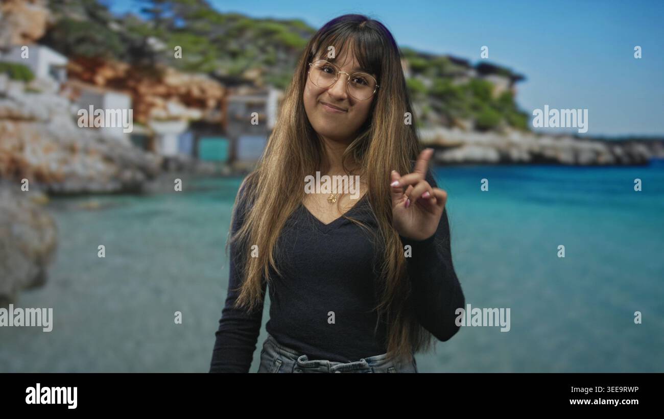 Jeune femme pointe l'index à la caméra sur la plage, portant des lunettes et du denim, cheveux longs visibles, souriant doucement ; confiance ludique. Banque D'Images