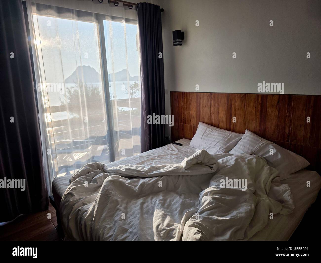 Paisible chambre du matin avec lit non fait froissé coussins de couverture blanche en bois tête de lit grande fenêtre laissant entrer la lumière du soleil créant une relaxation calme Banque D'Images