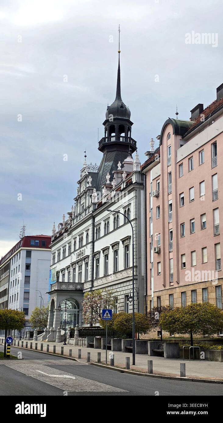 Un élégant bâtiment néo-renaissance avec une grande tour sombre se dresse à côté d'un immeuble rose le long d'une rue de la ville bordée de petits arbres Banque D'Images