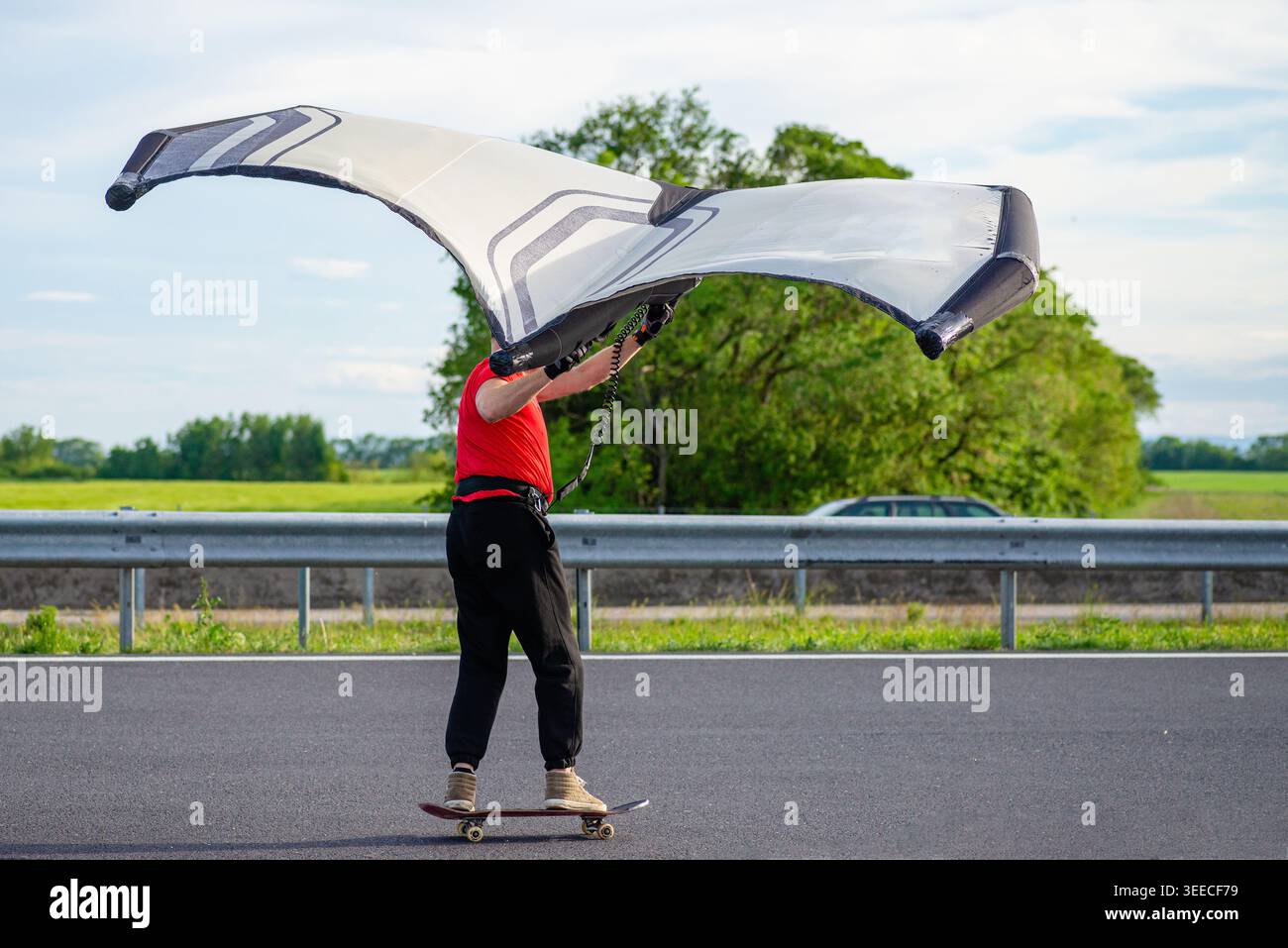 Vue latérale d'une personne sur un longboard tenant une aile foil pour attraper le vent sur une route ouverte pour l'entraînement de sports extrêmes Banque D'Images