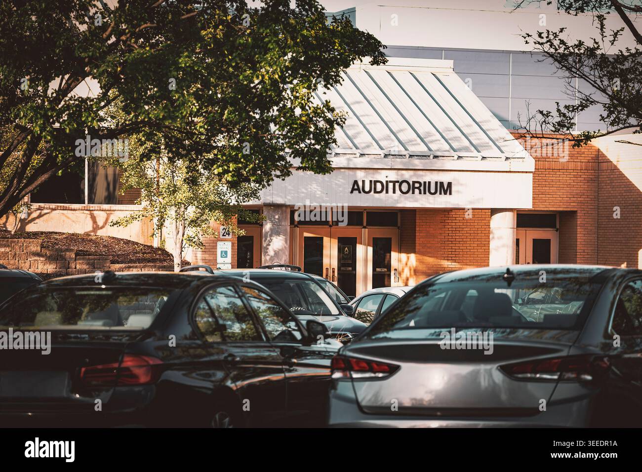 Entrée à un auditorium moderne en briques avec un toit à pignons blanc derrière un parking bondé avec des voitures et des arbres verdoyants à la lumière du jour Banque D'Images