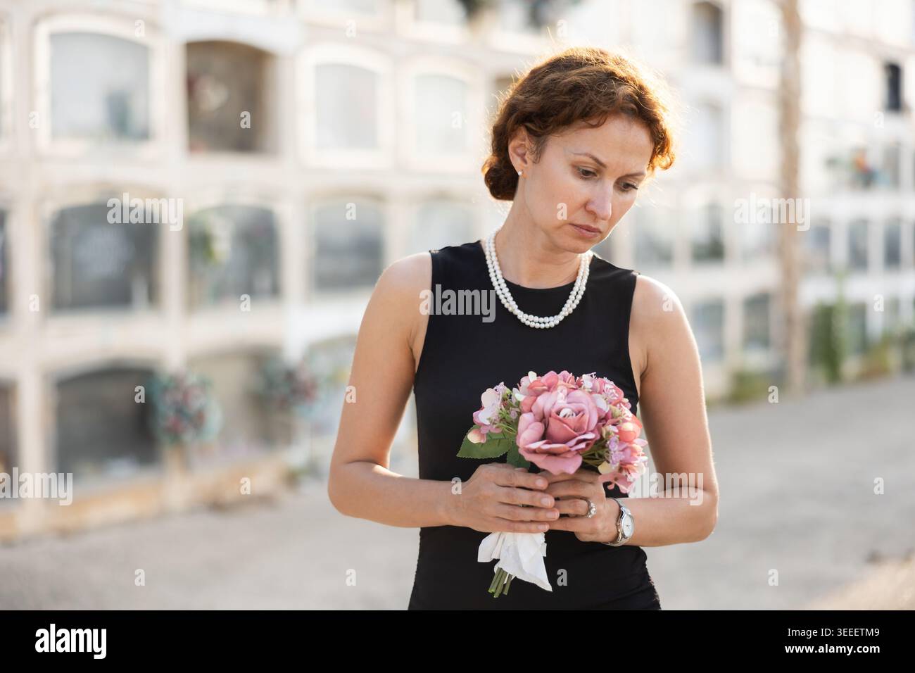 Femme triste en robe noire avec bouquet de fleurs et mouchoir dans les mains, se tient dans le cimetière Banque D'Images