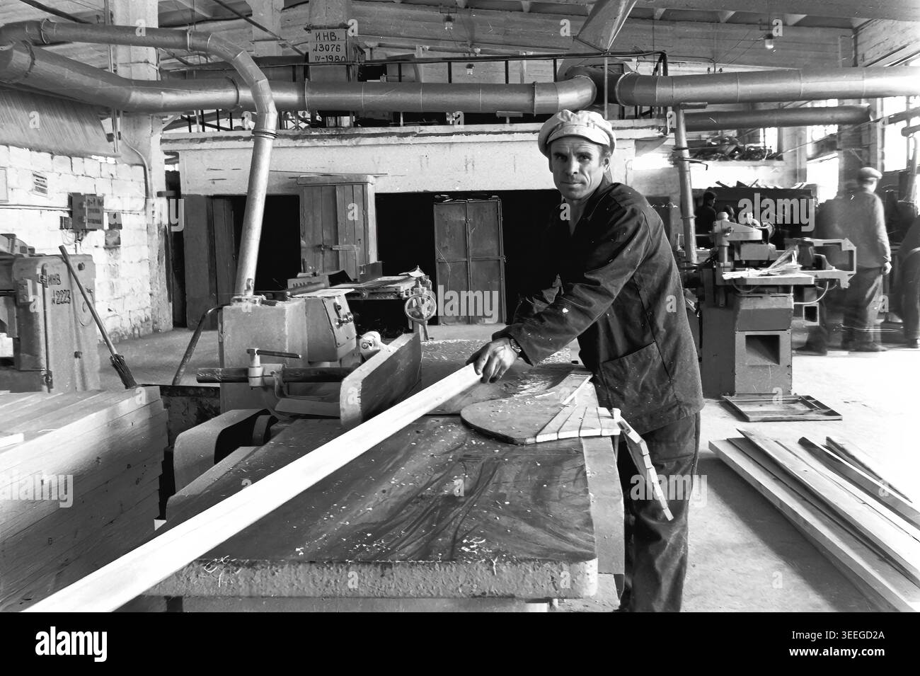 Slaviansk, 1983. À l'intérieur d'un atelier à l'armature-Insulator Plant (AIZ). Un ouvrier se tient devant une grande machine à travailler le bois, guidant soigneusement une longue planche de bois à travers la raboteuse tout en regardant directement la caméra. Cette photographie vintage en noir et blanc capture l'atmosphère authentique de l'époque, reflétant les activités quotidiennes et la culture industrielle de la région pendant la période soviétique. Il sert de document historique précieux de la communauté locale, préservant la mémoire visuelle du peuple, mettant en valeur un Donbass pacifique. Banque D'Images