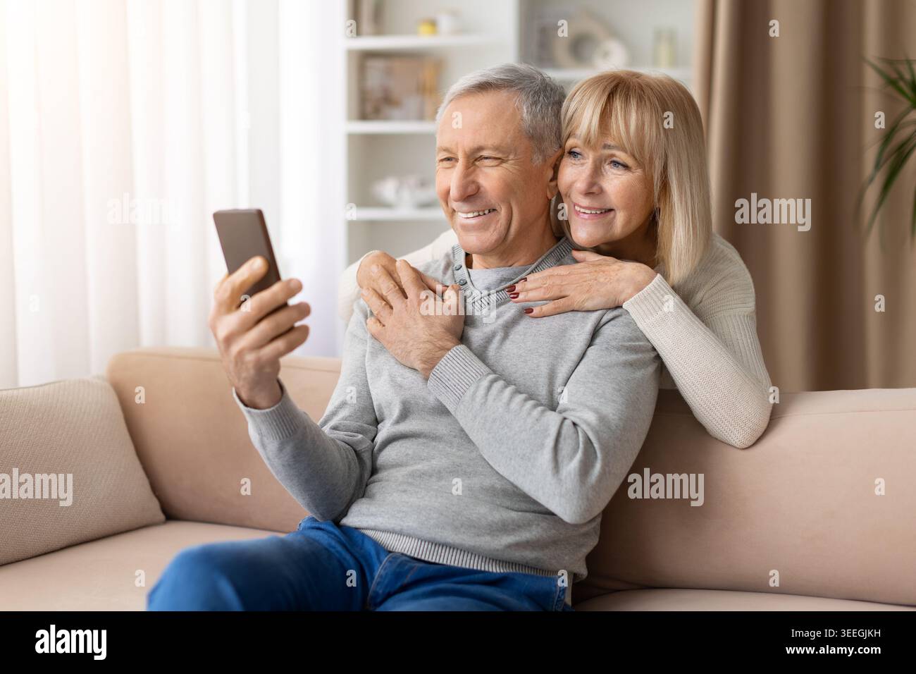 Amoureux couple mature embrasser tout en utilisant smartphone à la maison, profiter de la communication en ligne Banque D'Images
