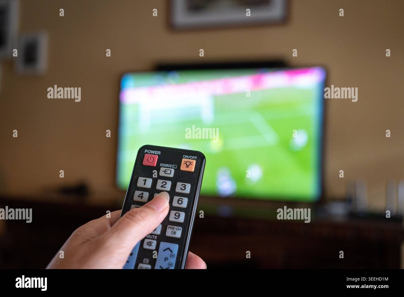 Regarder un match de football à la maison, se concentrer sur la main tenant la télécommande avec l'écran de télévision défocalisé du match de football en arrière-plan. Banque D'Images