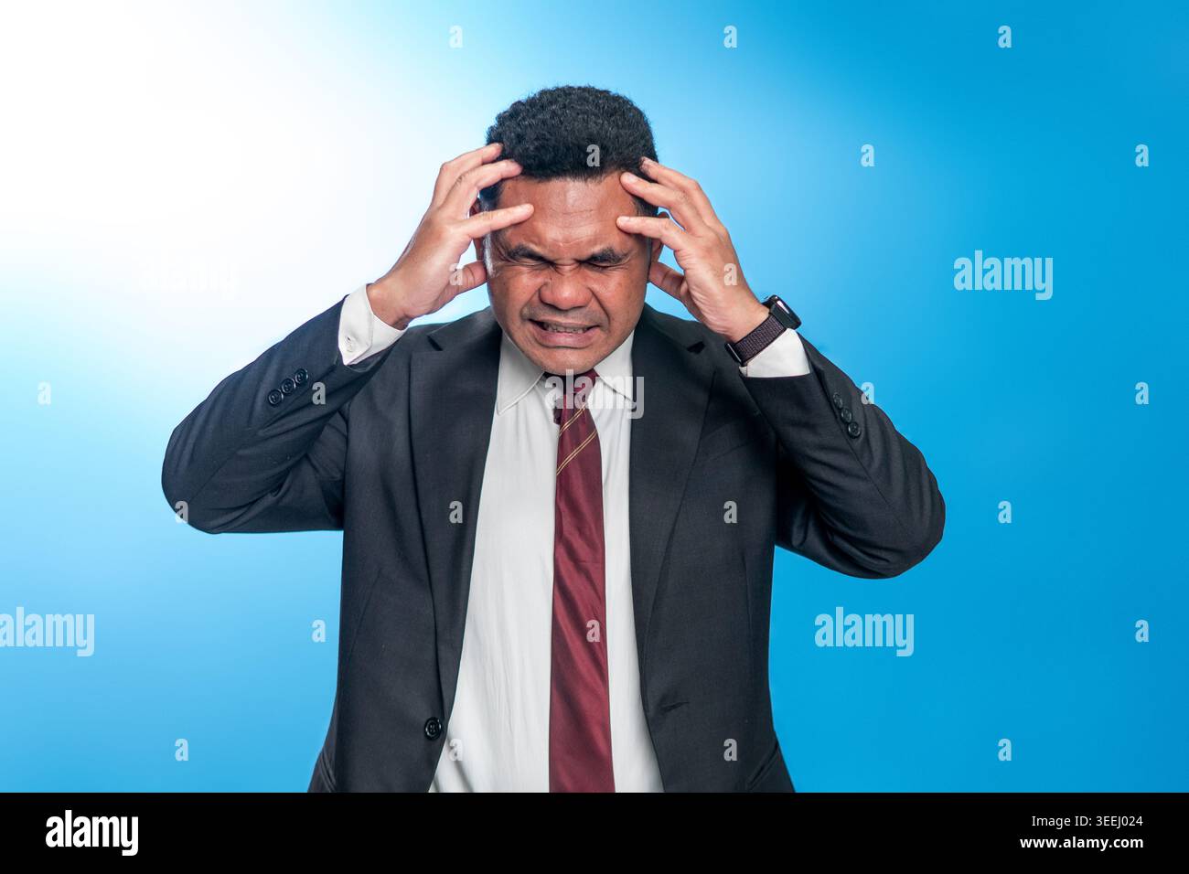 Portrait d'un homme indonésien stressé dans un costume tenant sa tête dans la douleur. Studio tourné sur fond bleu représentant le stress de l'entreprise ou la dette. Banque D'Images