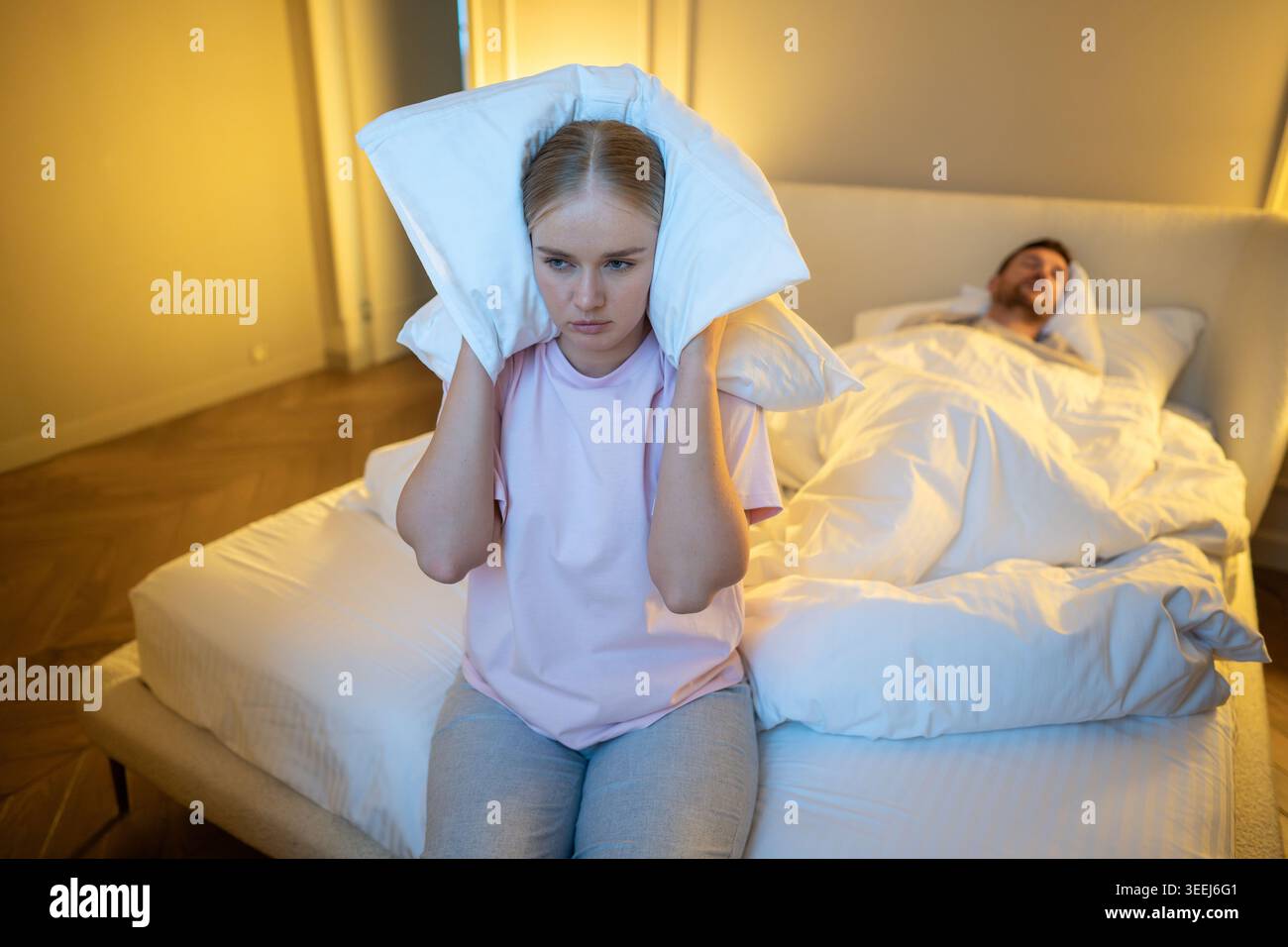 Épouse irritée assise sur le lit couvrant les oreilles avec un oreiller pour bloquer le bruit de ronflement pendant que l'homme dort. Banque D'Images