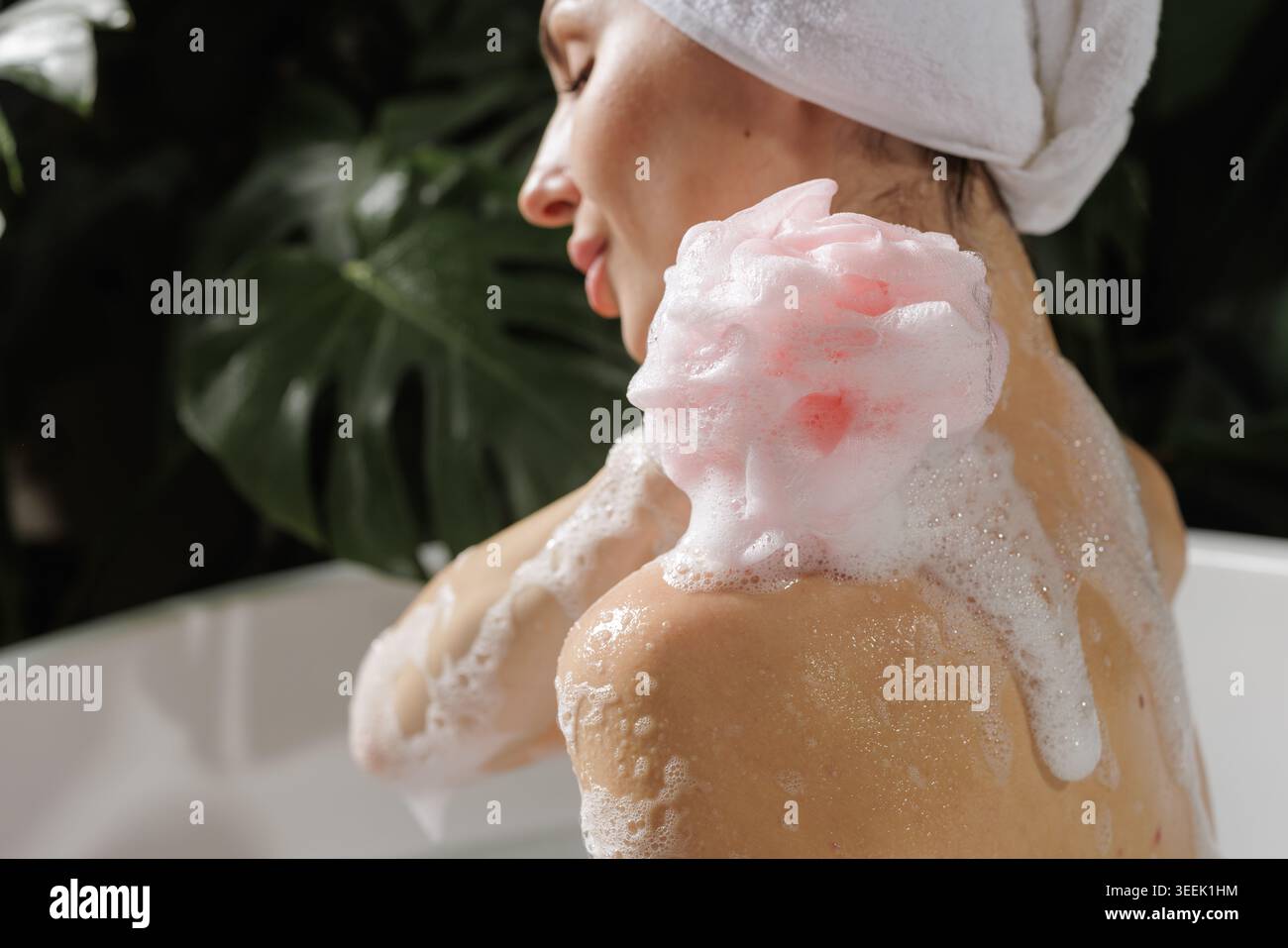 Une femme se détend dans une salle de bain de type spa, en appliquant du shampooing avec de la mousse sur son épaule, entourée d'une végétation luxuriante, parfaite pour le bien-être et la beauté campa Banque D'Images