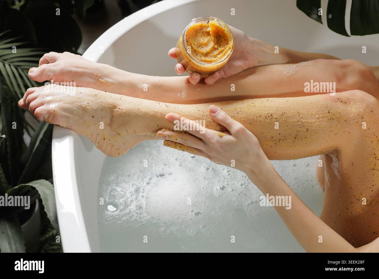 Une femme applique un gommage corporel naturel sur ses jambes tout en se relaxant dans une baignoire, favorisant l'auto-soin et le rajeunissement de la peau dans un environnement serein semblable à un spa Banque D'Images