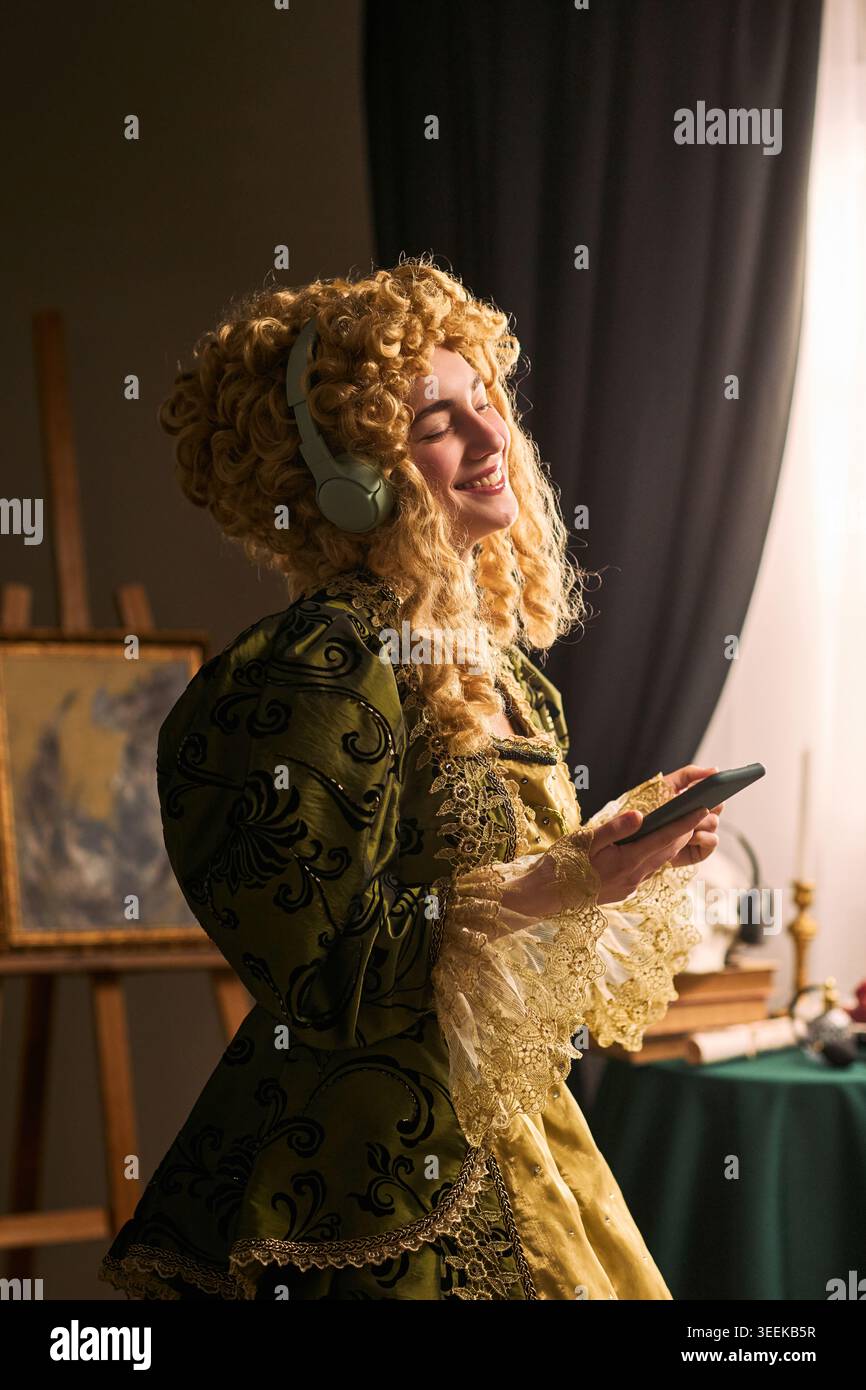 Photo verticale d'une jeune femme portant un costume historique, écoutant avec des écouteurs et utilisant un smartphone, mêlant culture patrimoniale et divertissement numérique. Pour le marketing des musées, la promotion des événements Banque D'Images