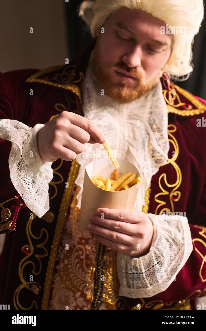 Plan vertical d'un jeune homme portant un costume historique, mangeant des frites dans un carton à emporter, mêlant humour et culture de consommation. Idéal pour les annonces fast food ou satire, vertical Banque D'Images