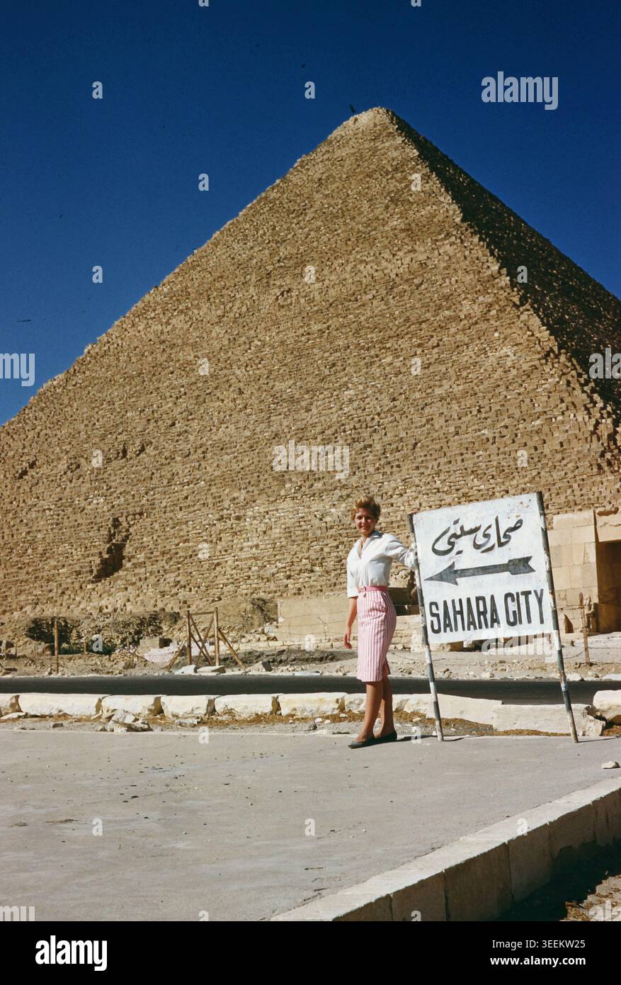Une touriste posant en plein air à côté d'un panneau directionnel en bois blanc abîmé indiquant "SAHARA CITY" en lettres noires épaisses en anglais et en écriture arabe manuscrite, encadré de près par les énormes gradins en blocs de calcaire d'une pyramide de Gizeh. Banque D'Images