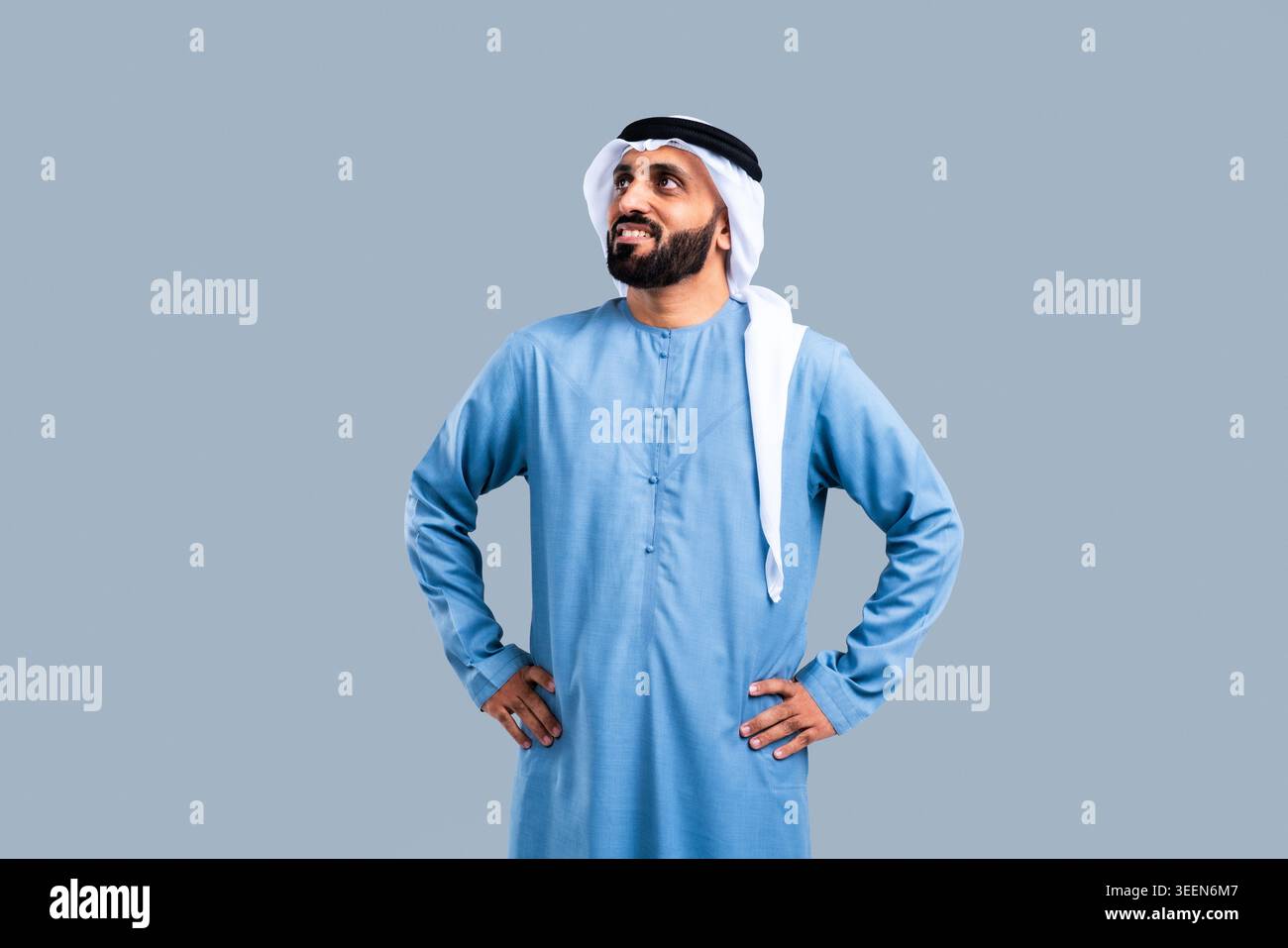 Homme émirati en kandura et ghutra traditionnels, les mains sur les hanches, souriant avec confiance et regardant vers le haut avec espoir, expression visionnaire contre une lueur Banque D'Images