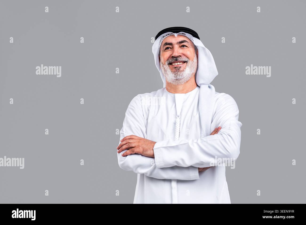 Homme émirati mature senior souriant, portant des kandora et ghutra traditionnels, levant les yeux avec les bras croisés, dépeignant la confiance, la sagesse et un optime Banque D'Images