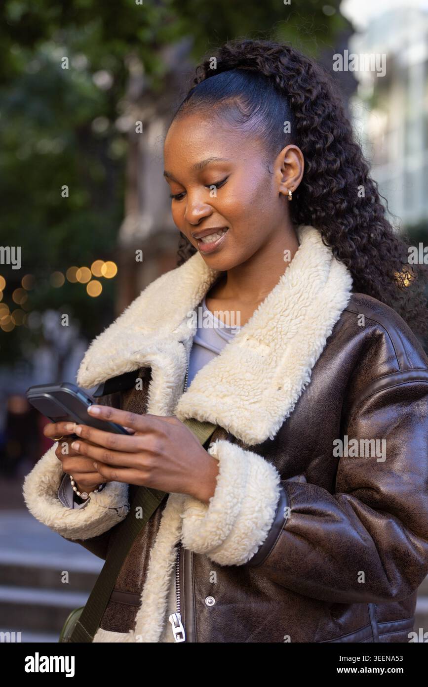 Femme africaine adulte debout dans la rue regardant vers le bas le smartphone portant une veste doublée de cisaillement Banque D'Images