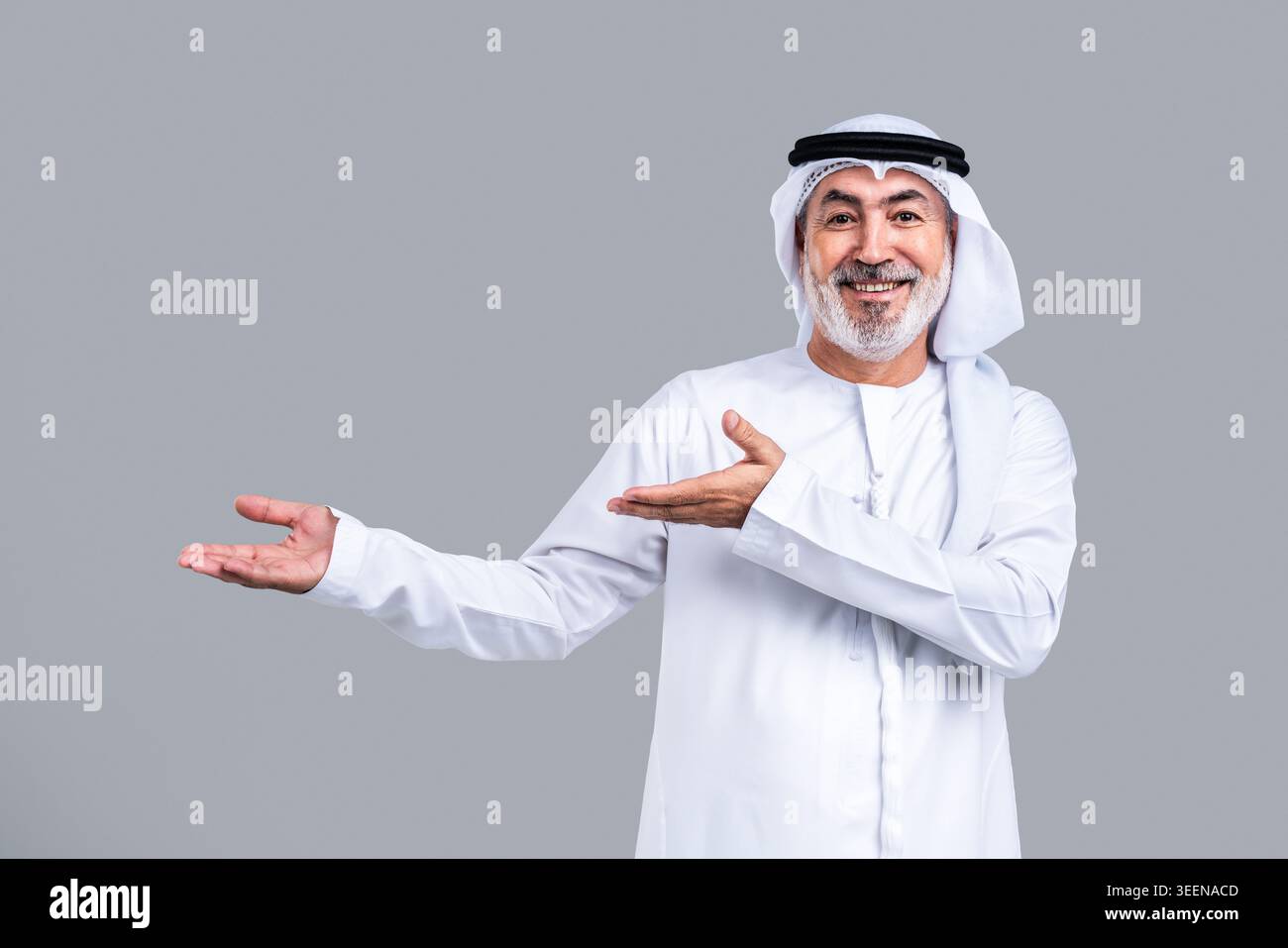Homme arabe mature senior en kandora, ghutra et agal émiratis traditionnels, souriant largement et faisant des gestes avec les mains ouvertes, indiquant un accueil et un pré Banque D'Images