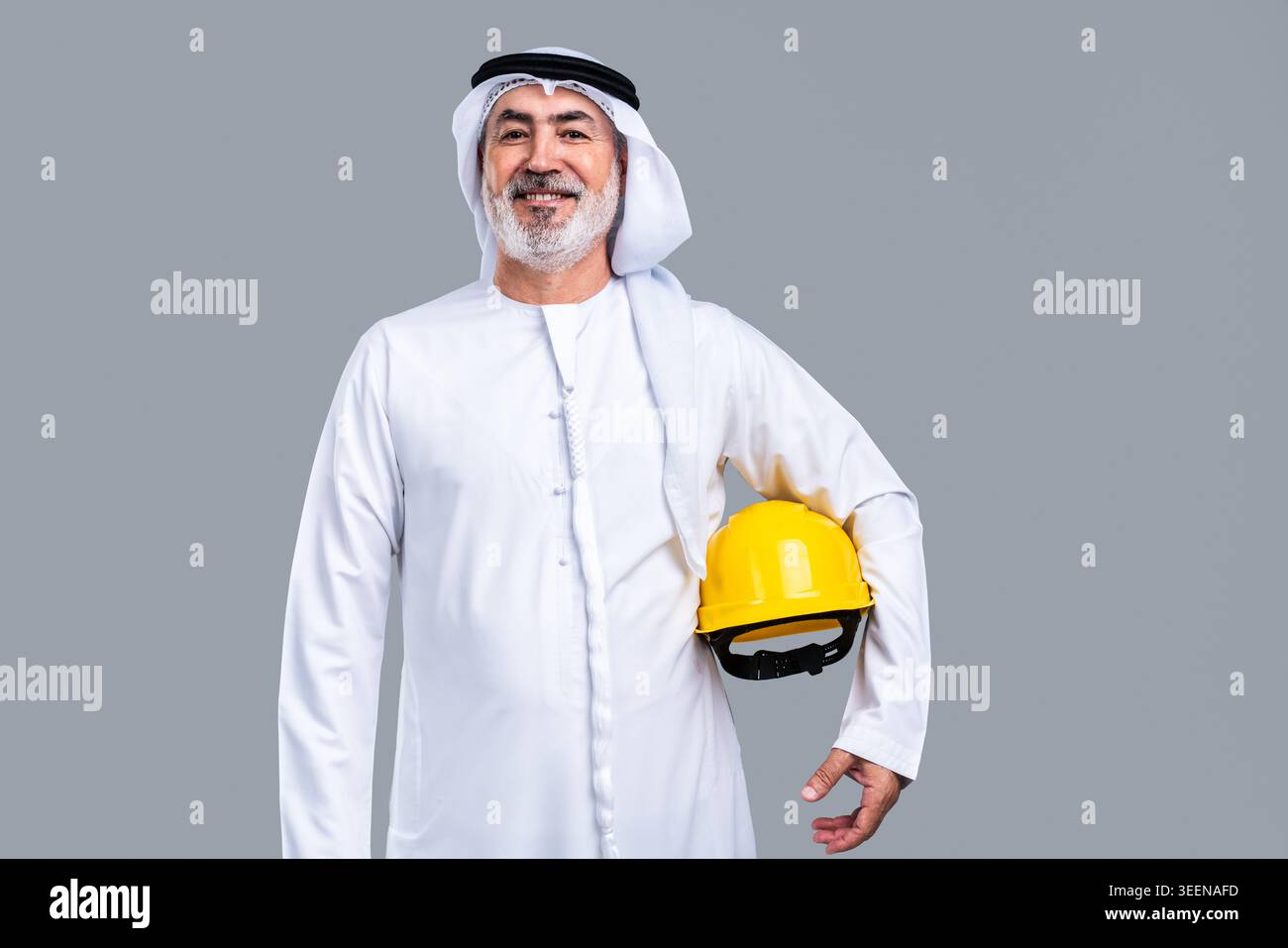 Homme émirati senior portant des kandora et ghutra traditionnels, souriant avec confiance tout en tenant un casque jaune, représentant l'expérience et la profession Banque D'Images