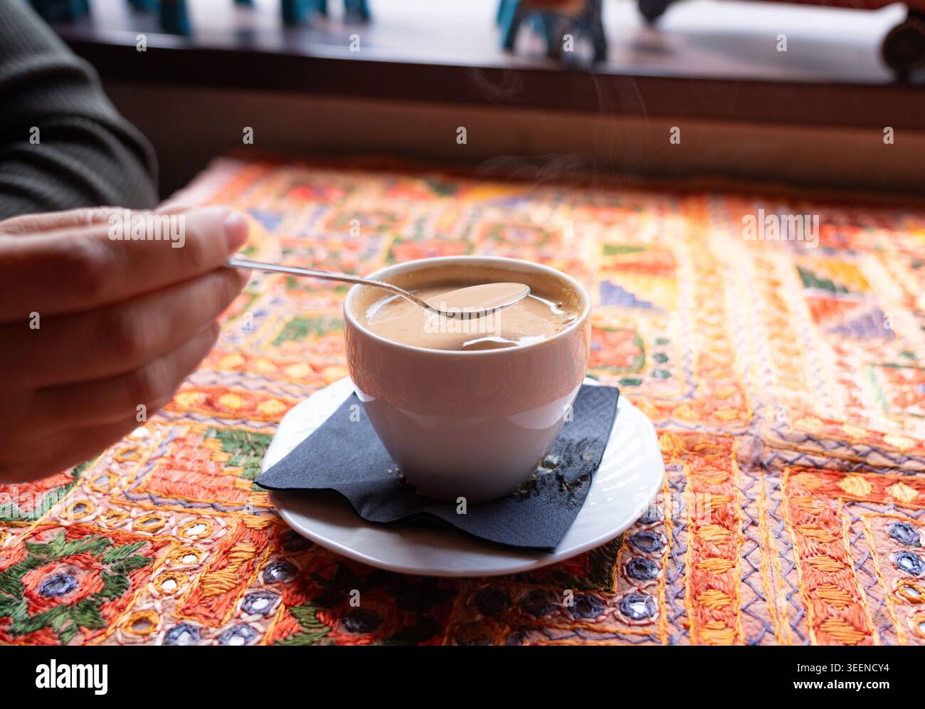 Une main remue le thé chaud ou le café avec du lait dans une tasse blanche sur une table avec un motif textile traditionnel sur fond flou, mise au point sélective Banque D'Images