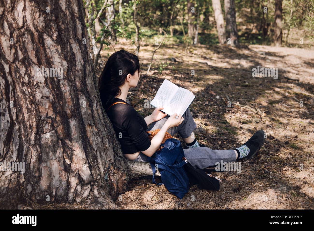 La jeune femme est assise contre le tronc d'arbre tout en lisant le livre dans la forêt. Sensibilisation à la santé mentale, pratique de pleine conscience, bien-être émotionnel, soulagement du stress Banque D'Images
