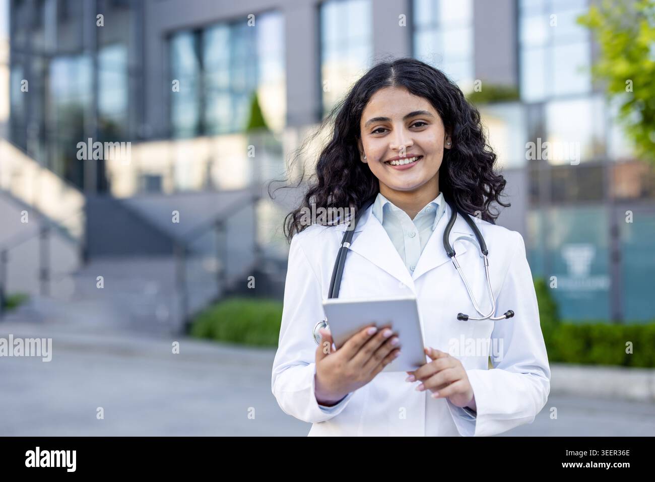 Jeune professionnel de la santé confiant portant une blouse blanche et un stéthoscope, utilisant une tablette tout en se tenant debout à l'extérieur d'un établissement de santé moderne, représentant l'innovation et le dévouement en médecine Banque D'Images