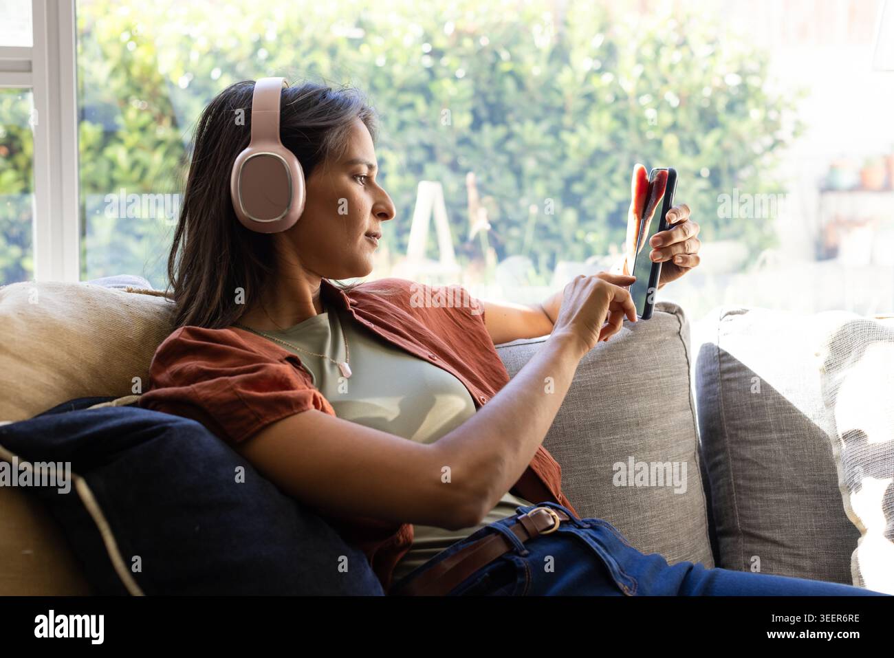 Femme indienne en vêtements décontractés couchée sur un canapé par fenêtre, utilisant un smartphone et portant des écouteurs Banque D'Images