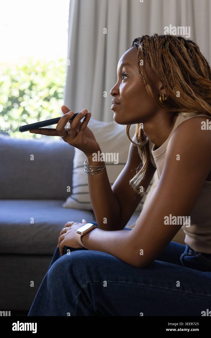 Femme africaine assise sur un canapé gris à la maison, tenant un appareil mince près de la bouche et parlant Banque D'Images